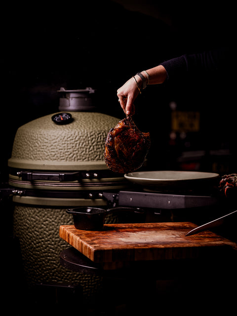 Person holding a seared steak over a large ceramic grill with a dark background