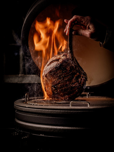 Hand holding a roasted steak over an open flame in a kamado oven.