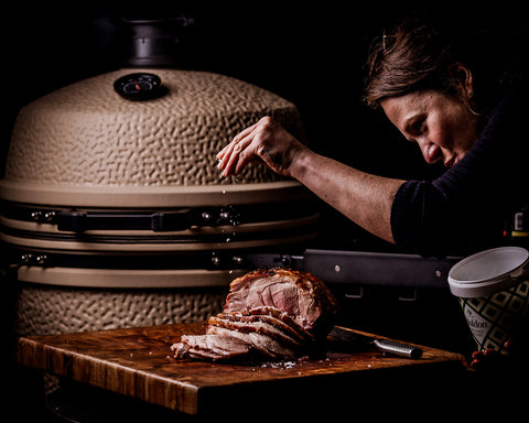 Person cutting a large piece of meat on a wooden board with a ceramic grill in the background.